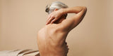 A nude woman touching her back against a beige background