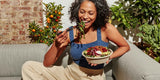 A woman sitting on a gray couch outdoors, smiling while eating a salad from a white bowl with a fork. She is wearing a blue ribbed tank top and beige pants, with plants and a brick wall in the background.
