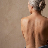 A menopausal woman's bare back with her grey hair tied into a bun, against a warm stone background