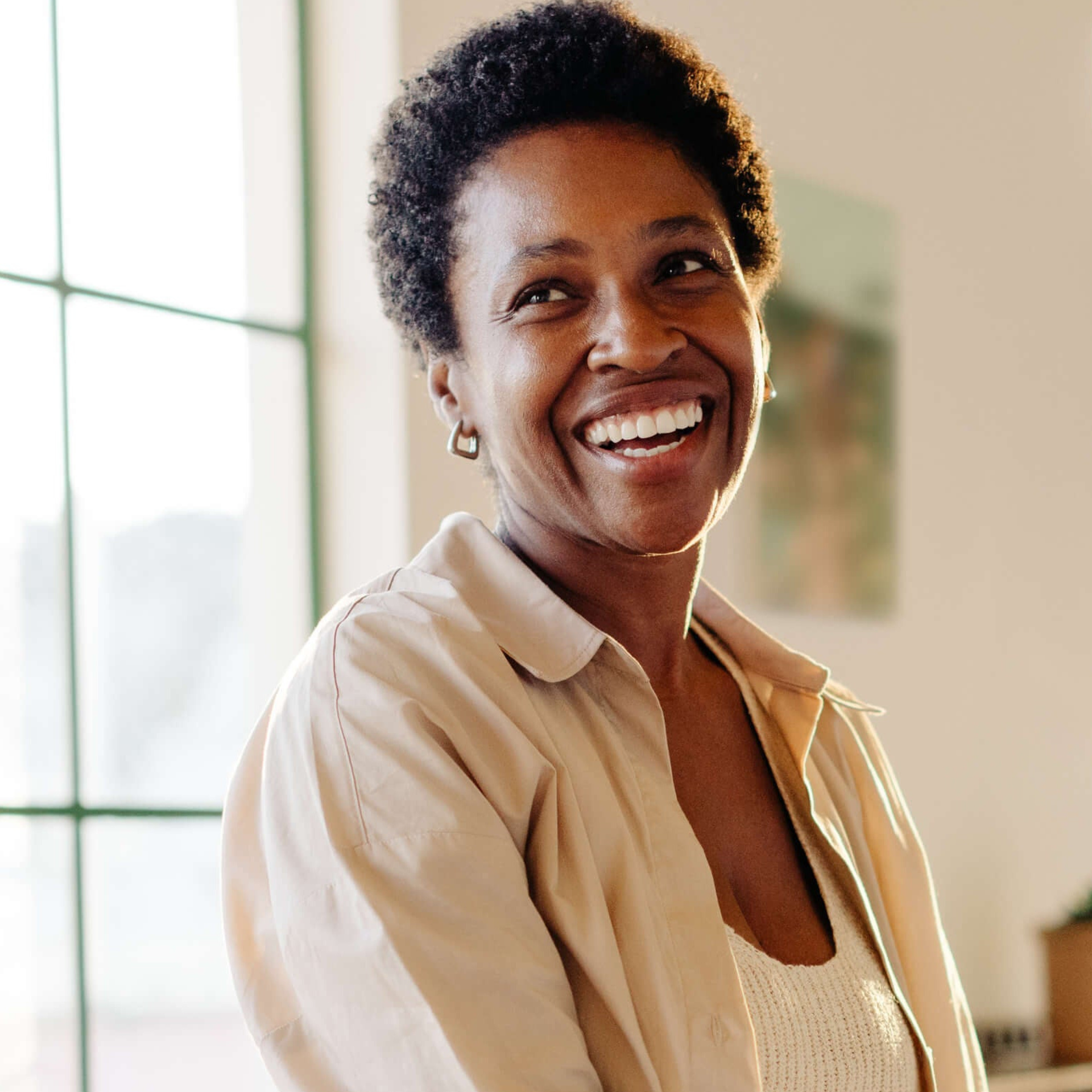 A smiling woman with short curly dark hair, wearing a beige button-up shirt over a white knit top, standing in a brightly lit indoor setting.