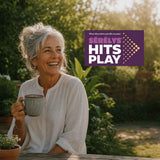 A woman in a garden holding a ceramic mug with a 'Serelys Hits Play' rectangular banner to the right of her