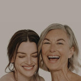A young woman and an older woman with their heads together and laughing against a neutral background