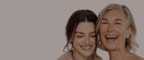 A young woman and older woman with their heads together and smiling against a neutral grey background